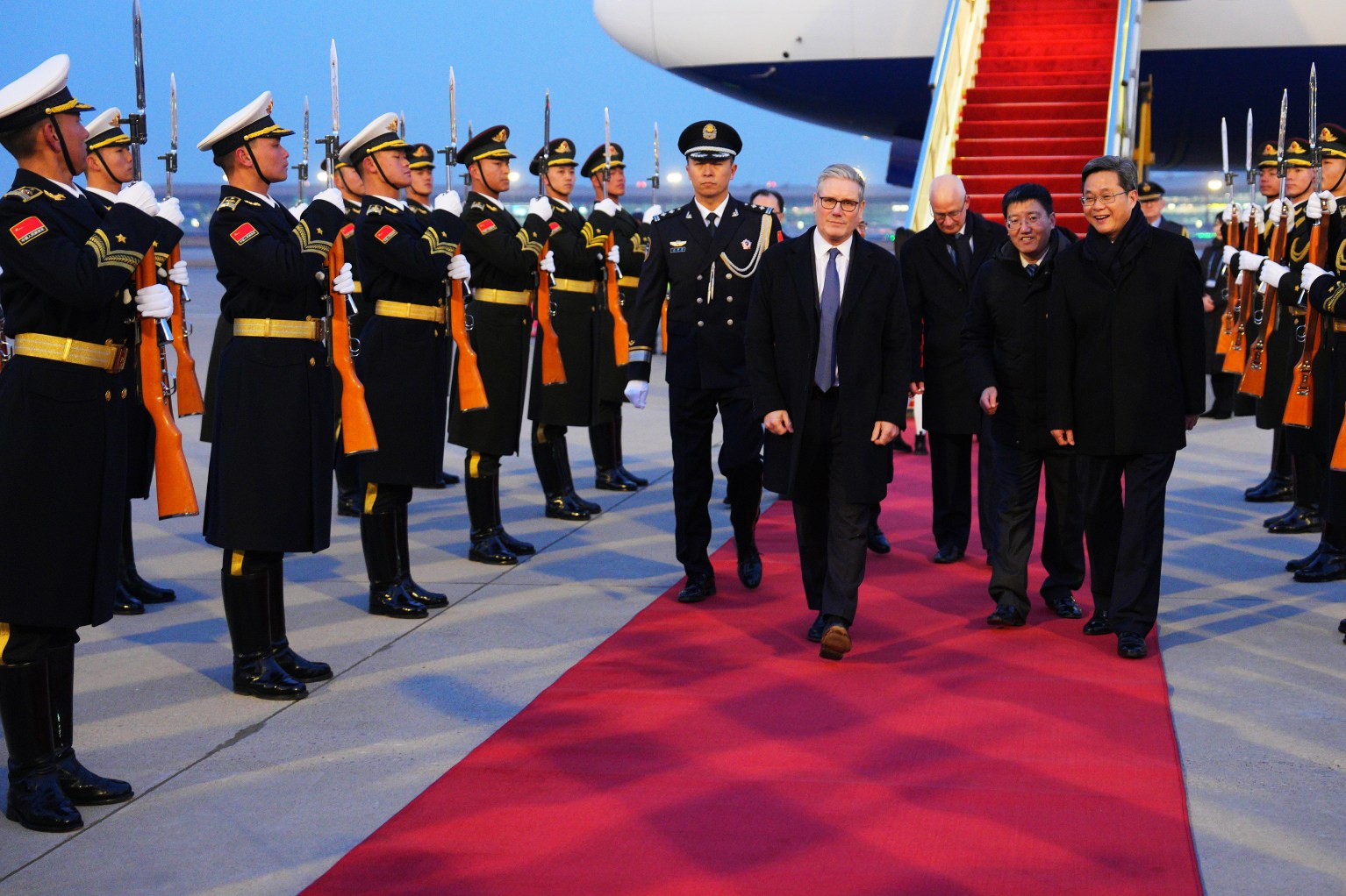 UK Prime Minister Keir Starmer walking on a red carpet after disembarking from a plane, with a military honor guard standing on the left.